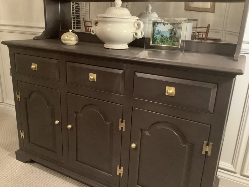 May include: A dark brown wooden hutch with three drawers and two doors. The hutch has brass hardware and a mirrored back.