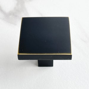 Op de afbeelding: Zwarte vierkante kastknop met een gouden rand. De knop heeft een vierkante basis.