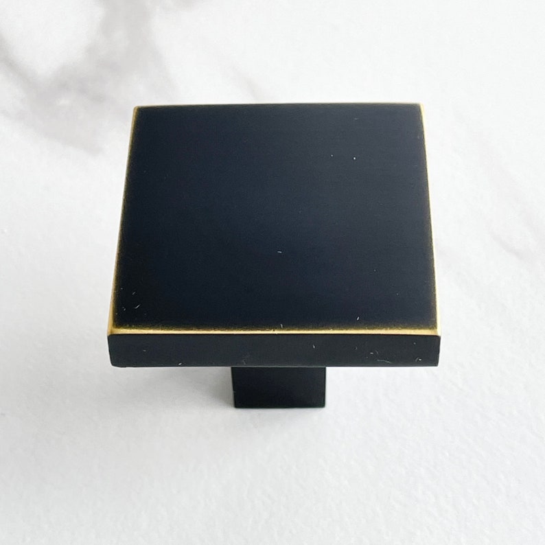 Op de afbeelding: Zwarte vierkante kastknop met een gouden rand. De knop heeft een vierkante basis.