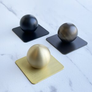 May include: Three round knobs in different finishes: black, brass, and gold. Each knob is mounted on a square base.