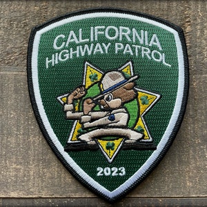May include: A green and yellow embroidered patch with the text "CALIFORNIA HIGHWAY PATROL" and a cartoon bear wearing a hat and holding a stop sign. The year 2023 is embroidered at the bottom of the patch.