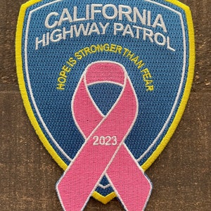 May include: A blue and yellow embroidered patch with the text "California Highway Patrol" and a pink ribbon with the text "Hope is Stronger Than Fear" and "2023".