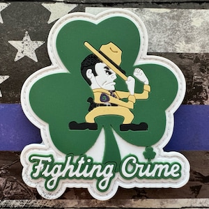 Può includere: Un cerotto a forma di trifoglio verde con un personaggio dei cartoni animati che indossa un cappello e tiene un bastone. Il testo "Fighting Crime" è scritto in bianco nella parte inferiore del cerotto.