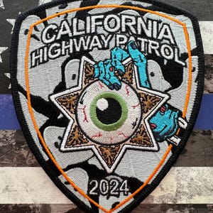 May include: A white and black embroidered patch with an orange border. The patch features a stylized eye with a green iris and a blue hand holding it. The text "CALIFORNIA HIGHWAY PATROL" is written in white on a black background. The year "2024" is written in orange at the bottom of the patch.