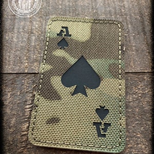 May include: A black and green camouflage fabric patch with a black ace of spades playing card design. The patch has a stitched border.