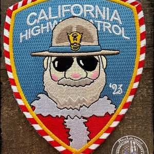 May include: Embroidered patch featuring a cartoon Santa Claus wearing a California Highway Patrol uniform and sunglasses. The patch is blue with a white border and red and white candy cane stripes around the edge. The text "CALIFORNIA HIGHWAY PATROL" is written in white on the patch. The year "'23" is written in white on the right side of the patch.