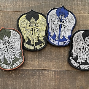 Puede incluir: Cuatro parches bordados con un ángel blanco con alas sosteniendo una espada. Cada parche tiene un color de fondo diferente: negro, azul, verde y amarillo. El texto "ST. MICHAEL PROTECTUS" está escrito en cada parche.