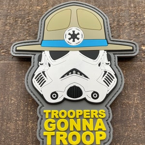 Könnte beinhalten: Ein grauer, weißer und gelber gestickter Aufnäher mit einem Cartoon-Bild eines Stormtroopers, der einen Khaki-Rangerhut mit einem blauen Band trägt und den Text "TROOPERS GONNA TROOP".