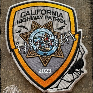 May include: Embroidered patch featuring the words "CALIFORNIA HIGHWAY PATROL" above a star-shaped emblem. The patch has a gray background with orange and black accents, and the year "2023" is displayed. A spider web design is on the right side.