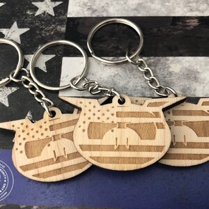 May include: Three wooden keychains with an American flag design. Each keychain features a stylized image of a helmet with wings. The keychains are attached to silver key rings.