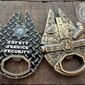Könnte beinhalten: Zwei goldfarbene Flaschenöffner in Form des Millennium Falcon aus Star Wars. Eine Seite hat ein schwarzes und silbernes Diamantplatten-Design mit den Worten "SAFETY SERVICE SECURITY" und einem Logo der California Highway Patrol. Die andere Seite zeigt ein detailliertes Bild des Millennium Falcon.