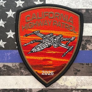 May include: A shield-shaped patch with the text "CALIFORNIA HIGHWAY PATROL" in orange embroidery. The patch features a detailed silver spaceship design over a red and orange sunset background, with the number "2026" at the bottom.
