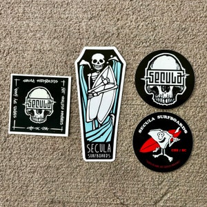 May include: Three black and white stickers with a skull motif. The stickers feature the text "Secula Surfboards" and "Shaded by Hand, Not Soulless Machines".