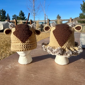 May include: Two handmade crochet hats in the shape of a cow's head. The hats are tan with brown accents, including a diamond-shaped patch on the forehead and small ear details. One hat has a faux fur trim around the base.