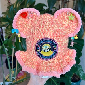 Könnte beinhalten: Ein handgefertigter, rosa und gelber gehäkelter Hut mit einem einzigartigen Design. Der Hut hat einen runden Aufnäher mit dem Text "SUBTRONICS CYCLOPS ARMY" und verschiedenen Anhängern. Der Hut hat einen gerüschten Rand und ohrähnliche Vorsprünge.