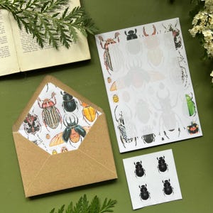 以下が含まれることがあります： 茶色の封筒、メモ帳、ステッカーシートがセットになった文房具セット。すべてカラフルな昆虫のイラストで飾られています。封筒の内側とメモ帳の縁には、さまざまなカブトムシが描かれています。ステッカーには黒いカブトムシのデザインが施されています。