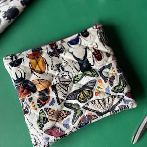 Könnte beinhalten: Eine cremefarbene, gesteppte Tasche mit einem lebendigen Insekten- und Schmetterlingsdruck. Das Design umfasst verschiedene Käfer und Schmetterlinge in Rot-, Gelb-, Grün- und Blautönen. Die Tasche ist rechteckig mit einem Reißverschluss.