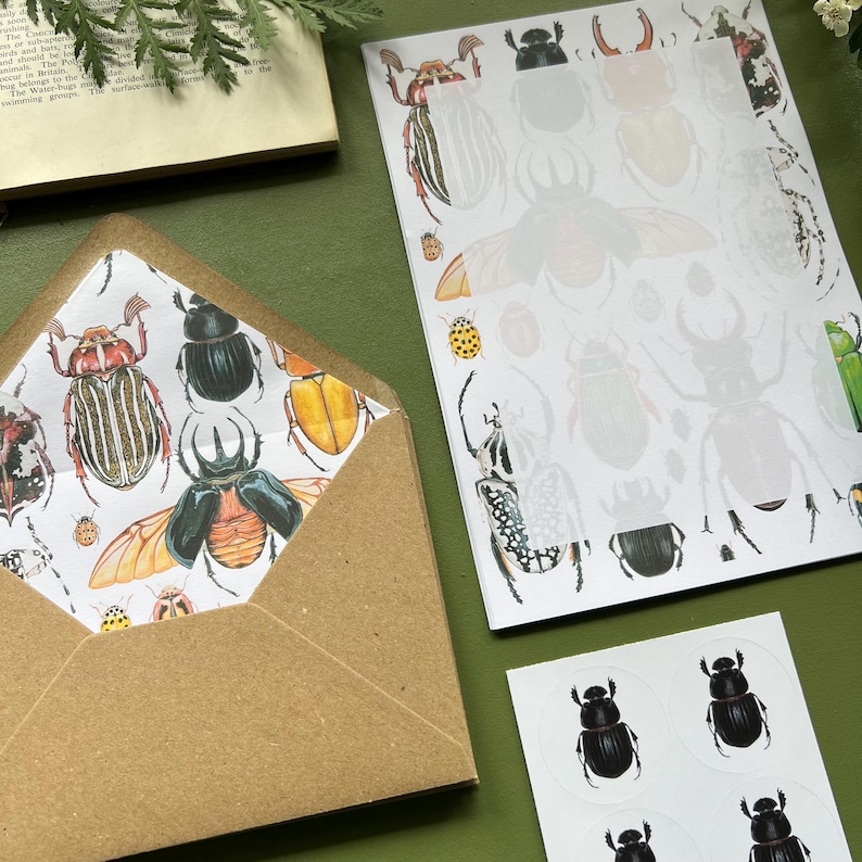 以下が含まれることがあります： カラフルなカブトムシのイラストが描かれた模様の茶色の封筒。カブトムシの輪郭が描かれたお揃いの紙と、黒いカブトムシのステッカーシートも付いています。アイテムは緑色の表面に配置されています。