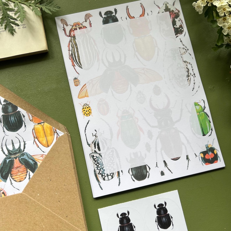 以下が含まれることがあります： 昆虫のイラストが特徴の文房具セット。セットには、落ち着いた色のカブトムシのデザインが施されたメモ帳、同様の昆虫アートが施された封筒、カブトムシのグラフィックが施されたステッカーシートが含まれています。