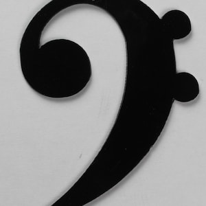 Könnte beinhalten: Schwarzer Bass-Schlüssel-Anhänger aus Metall. Der Anhänger ist in Form eines Bass-Schlüssels geformt und hat eine glatte, matte Oberfläche.