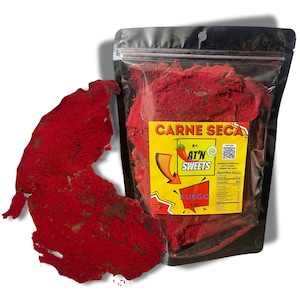 May include: A package of red Carne Seca, with a piece of the product displayed next to the package. The package has a yellow label with the text "CARNE SECA" and "AT'N SWEETS". The product is labeled "FUEGO" and is 4 oz.