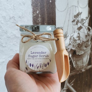 May include: A glass jar filled with a white sugar scrub with a brown twine bow and a wooden scoop. The jar has a label that reads "Cozy Bath and Body Co. Lavender Sugar Scrub Ingredients: Sugar, organic lavender essential oil & olive oil Net Wt 5 oz."
