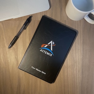 May include: Black leather notebook with the Artemis logo and the text "Your Name Here".