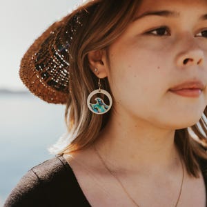 May include: A woman wearing a straw hat with a blue and white wave design earrings. She is also wearing a black shirt and a gold necklace with a pearl pendant.