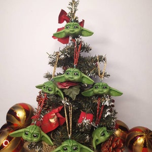 Puede incluir: Un pequeño árbol de Navidad artificial decorado con adornos verdes y marrones con forma del personaje Baby Yoda de la serie Star Wars. El árbol está rodeado de adornos rayados dorados y rojos.