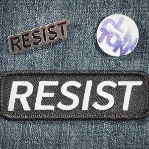 Puede incluir: Un surtido de accesorios con temática de protesta, que incluyen un pin de metal plateado y un parche en blanco y negro, ambos con la palabra "RESIST". También hay un botón redondo con la palabra "CLOWN" en morado.
