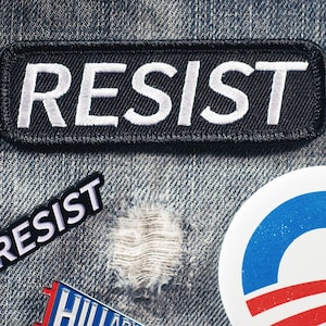 Puede incluir: Primer plano de una tela vaquera con varios parches y alfileres. El parche principal es negro con texto bordado en blanco que dice "RESIST". Los alfileres adicionales incluyen la palabra "RESIST" y un botón de campaña política.