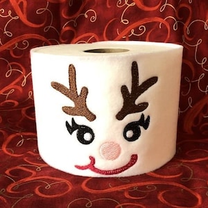 May include: White toilet paper roll with a red-nosed reindeer face embroidered on it. The reindeer has brown antlers, black eyes, and a pink nose.
