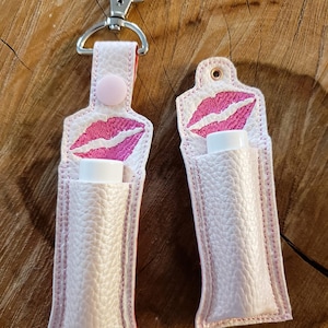 May include: Two white leather lip balm holders with pink stitching and a pink embroidered lip design. The holder on the left has a silver clip attached.