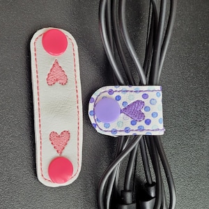 May include: Two white leather cord organizers with pink hearts and pink buttons. One organizer has a blue and white polka dot pattern with a purple button and a pink heart.
