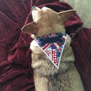 Può includere: Un piccolo cane marrone chiaro che indossa un bandana rosso, bianco e blu con una toppa a forma di osso. Il bandana ha un design patchwork con stelle e strisce.