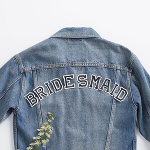 May include: A light-wash denim jacket with the word "BRIDESMAID" in black and white lettering across the back. A sprig of white and yellow flowers is placed on the left side of the jacket.