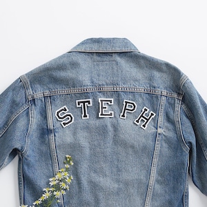 May include: A blue denim jacket with the name "STEPH" spelled out in black felt letters on the back.