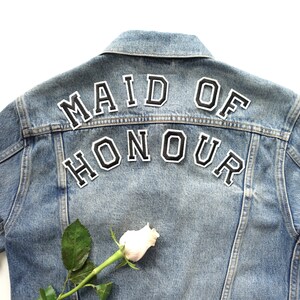 May include: A blue denim jacket with the words "Maid of Honour" in black letters on the back. A single white rose is on the bottom left side of the jacket.