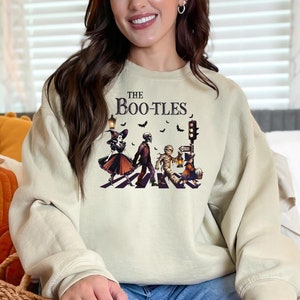 May include: Beige sweatshirt with the text "The BOO-TLES" and a graphic of Halloween characters crossing a street. The graphic includes a witch, a mummy, and a zombie. The sweatshirt is a comfortable, casual style.