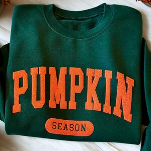May include: A dark green sweatshirt with the words "PUMPKIN SEASON" in orange block letters. The sweatshirt is folded on a white wooden surface, with fall-themed decorations in the background.