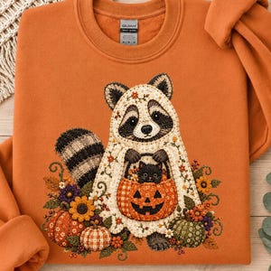 May include: An orange crewneck sweatshirt featuring a graphic of a raccoon wearing a white ghost sheet, holding a jack-o'-lantern bucket with a small black cat inside. The design includes autumn flowers, pumpkins, and a plaid pumpkin pattern.