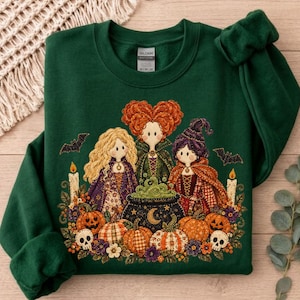 May include: A dark green crewneck sweatshirt featuring a graphic illustration of three stylized figures with long hair, wearing capes and dresses, standing around a bubbling cauldron with pumpkins, skulls, candles, and bats on a wooden surface.