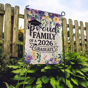 May include: A garden flag with the text "Proud Family of a 2026 Graduate" in black script and a graduation cap graphic. The flag is decorated with floral patterns in purple, yellow, and orange, and is displayed on a black metal stand.
