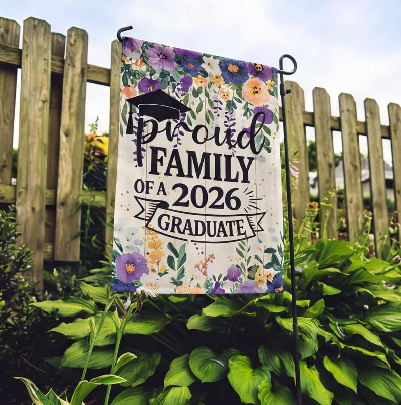 May include: A garden flag with the text "Proud Family of a 2026 Graduate" in black script and a graduation cap graphic. The flag is decorated with floral patterns in purple, yellow, and orange, and is displayed on a black metal stand.