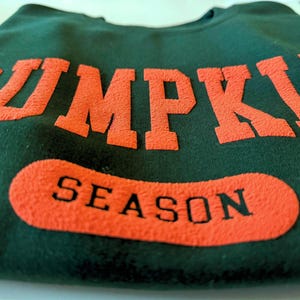 May include: Dark green sweatshirt with the words "PUMPKIN" in large orange block letters across the top and "SEASON" in a matching orange oval below. The sweatshirt is folded.