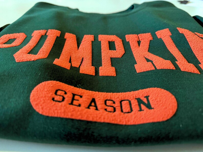 May include: Dark green sweatshirt with the words "PUMPKIN" in large orange block letters across the top and "SEASON" in a matching orange oval below. The sweatshirt is folded.