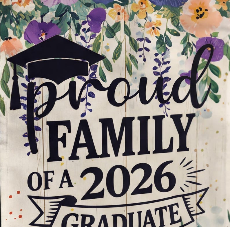 May include: A graduation-themed sign with the words "Proud Family of a 2026 Graduate" in black script. The sign features a graduation cap and is decorated with floral elements in shades of purple, orange, and yellow. The background has a wood-like texture.