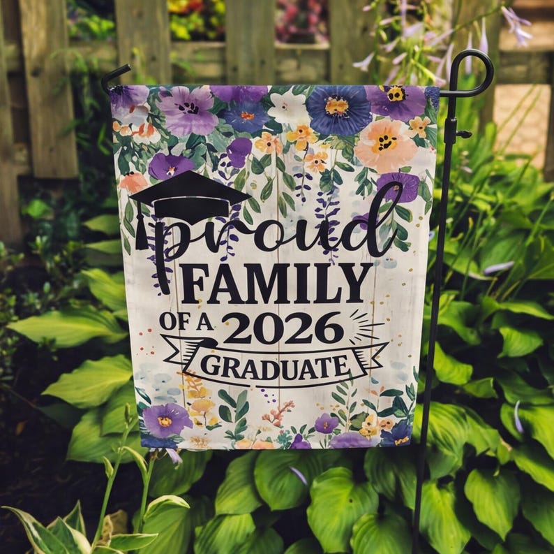 May include: A garden flag with a floral border in shades of purple, blue, and peach. The flag features the text "Proud Family of a 2026 Graduate" in black, with a graduation cap graphic. The flag is displayed on a black metal stand.