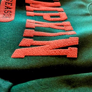 May include: Close-up of a dark green sweatshirt with the word "WAMP" in textured, red lettering. The word "SEASON" is also visible in red on the left side. The fabric has a soft, velvety texture.