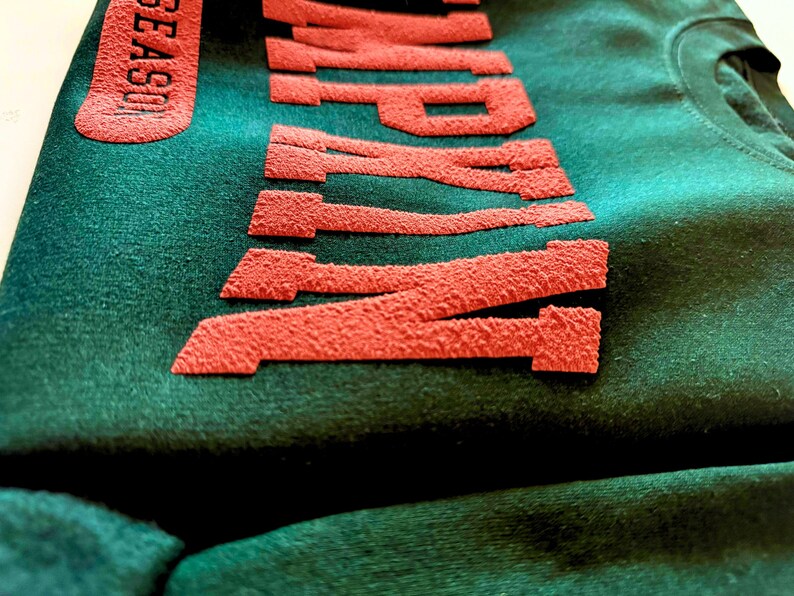 May include: Close-up of a dark green sweatshirt with the word "WAMP" in textured, red lettering. The word "SEASON" is also visible in red on the left side. The fabric has a soft, velvety texture.